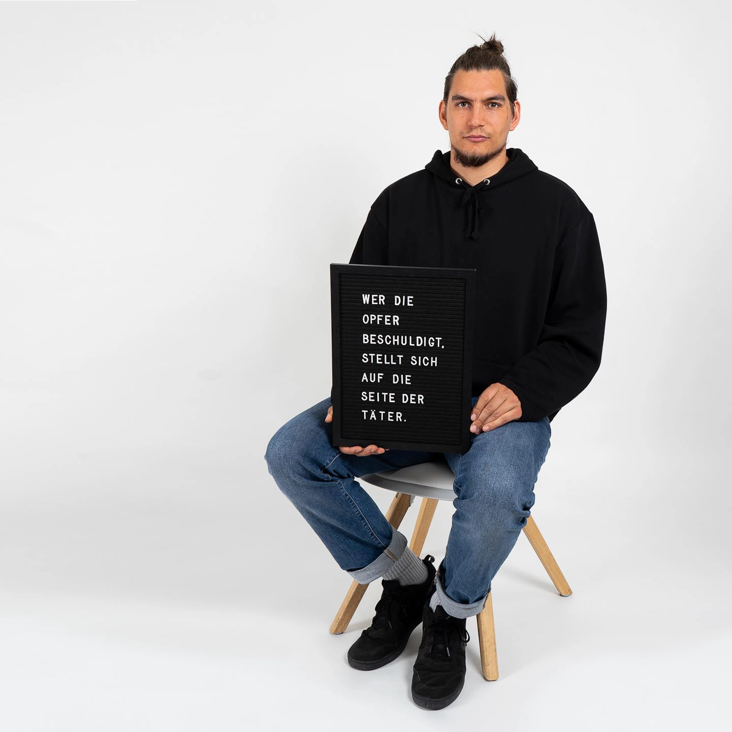 Ein Mann in schwarzem Pullover und Jeans sitzt auf einem Hocker und hält ein schwarzes Schild mit weißer Aufschrift für das Fotoshooting für 'Was hattest du an?'.