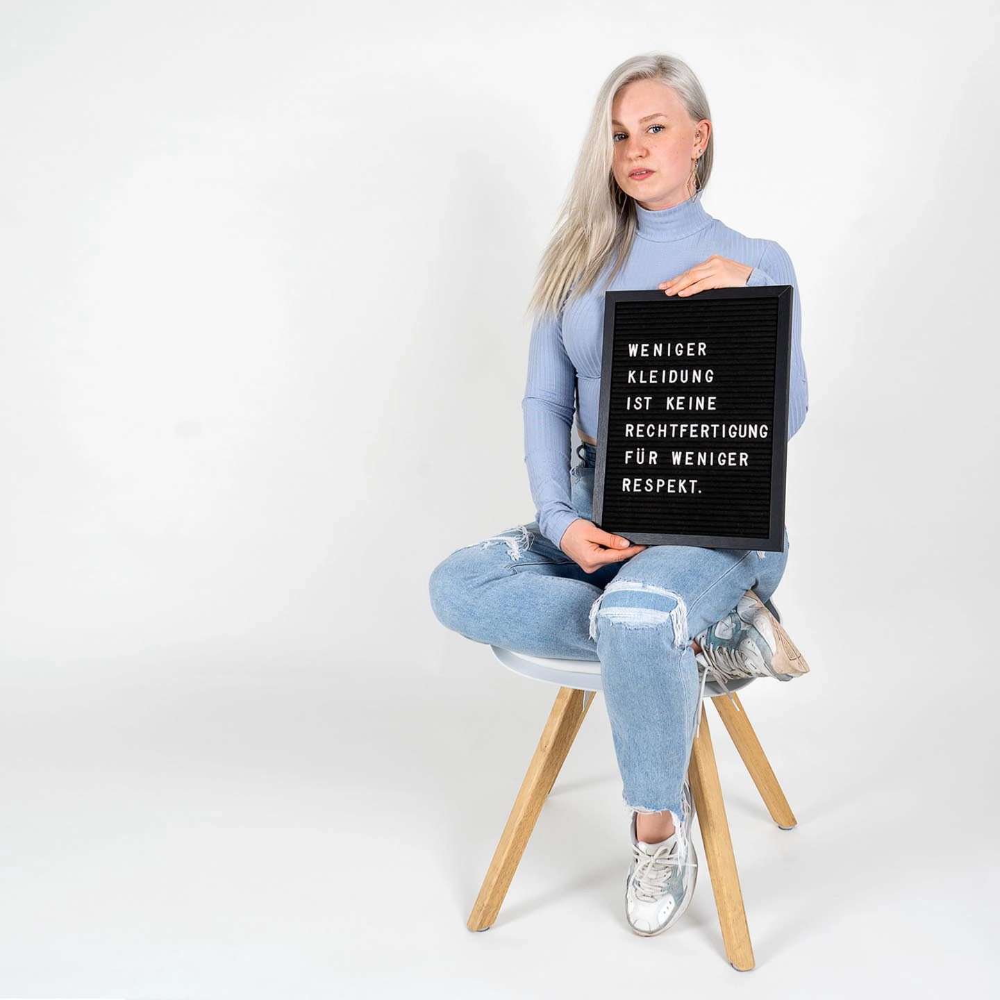 Eine Frau in blauem Pullover und Jeans sitzt auf einem Hocker und hält ein schwarzes Schild mit weißer Aufschrift für das Fotoshooting für 'Was hattest du an?'.