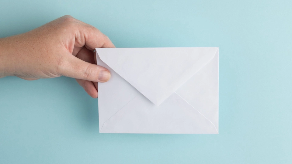 Hand hält einen weißen Briefumschlag vor einem blauen Hintergrund, symbolisch für E-Mail-Kommunikation.