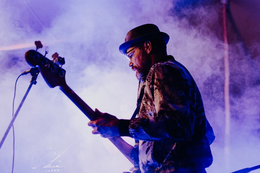 Musiker in einem bedruckten Hemd spielt Bassgitarre auf der Bühne beim Festungssommer mit blauem Licht und Nebel.