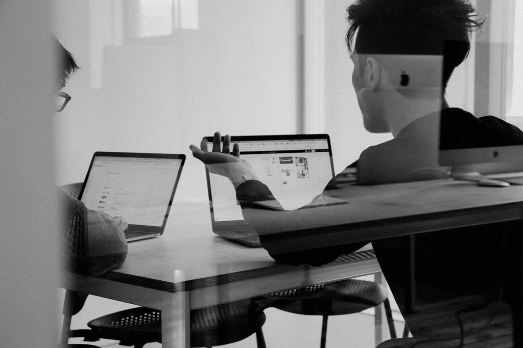 An einem Schreibtisch in einem hellen Büro diskutieren zwei Personen an ihren Laptops über Gestaltung und Fotodesign.