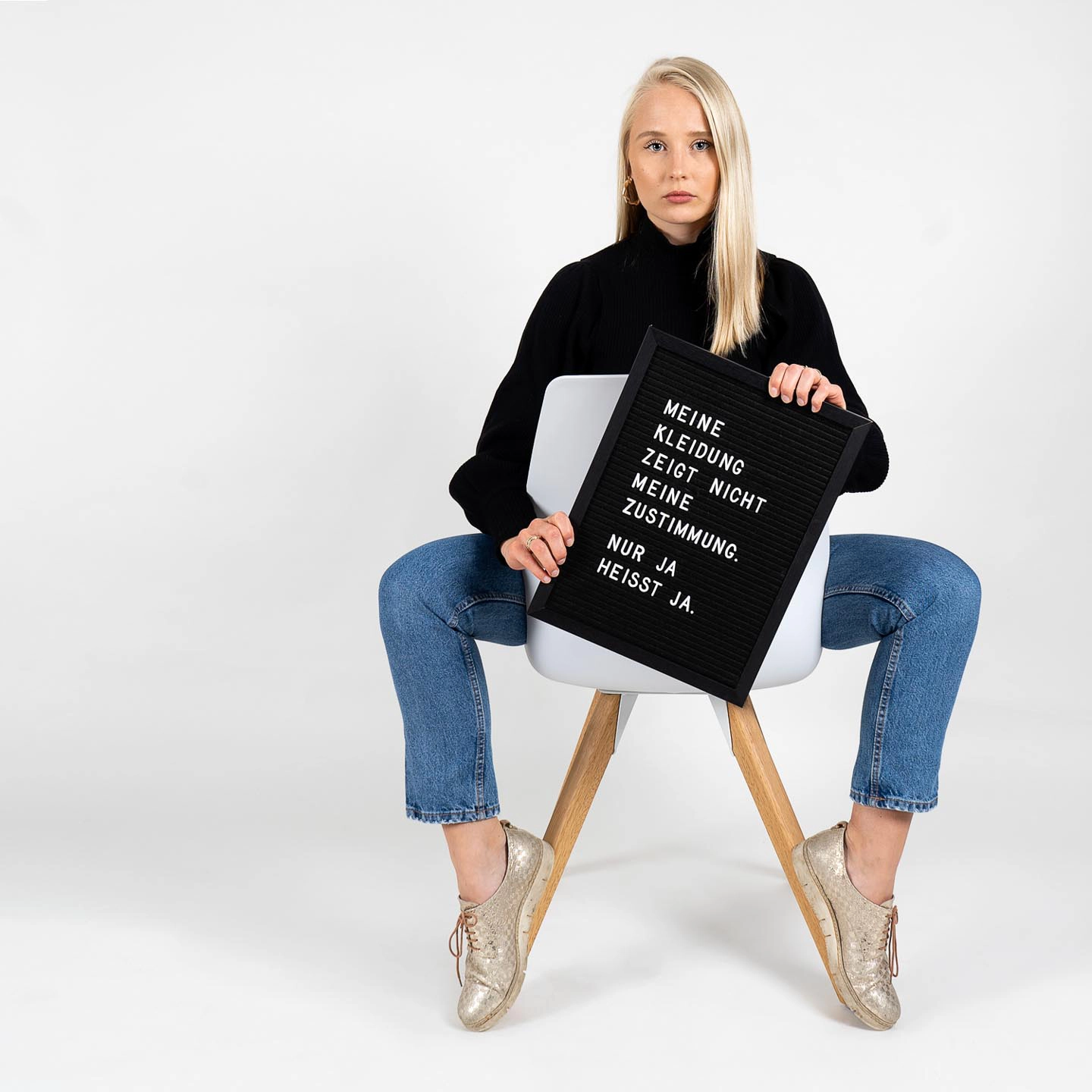 Eine Frau in schwarzem Pullover und Jeans sitzt auf einem Hocker und hält ein schwarzes Schild mit weißer Aufschrift für das Fotoshooting für 'Was hattest du an?'.