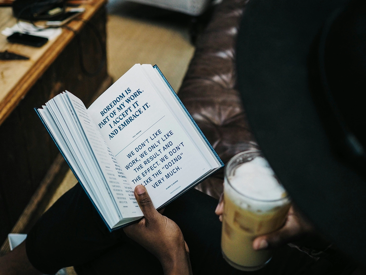 Eine Person liest ein Buch mit sichtbarem Text über Langeweile als Teil der Arbeit, während sie ein Glas mit einem cremigen Getränk hält.