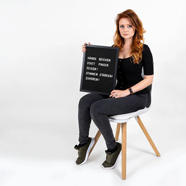 Eine Frau in schwarzem T-Shirt und Jeans sitzt auf einem Hocker und hält ein schwarzes Schild mit weißer Aufschrift für das Fotoshooting für 'Was hattest du an?'.
