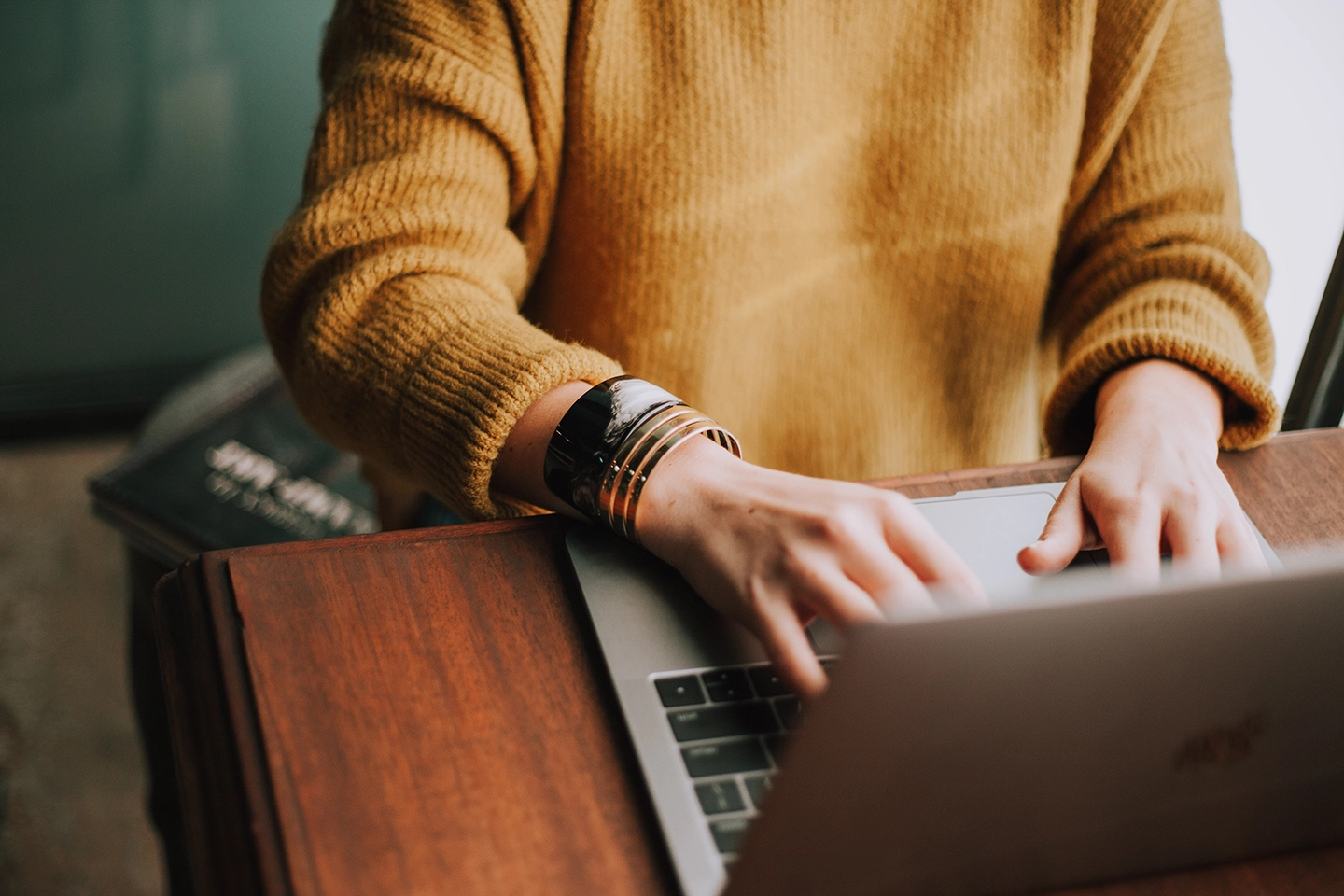 Eine Person in einem gelben Pullover tippt auf einem Laptop, der auf einem Holztisch steht und schreibt ihr Manuskript in Word.