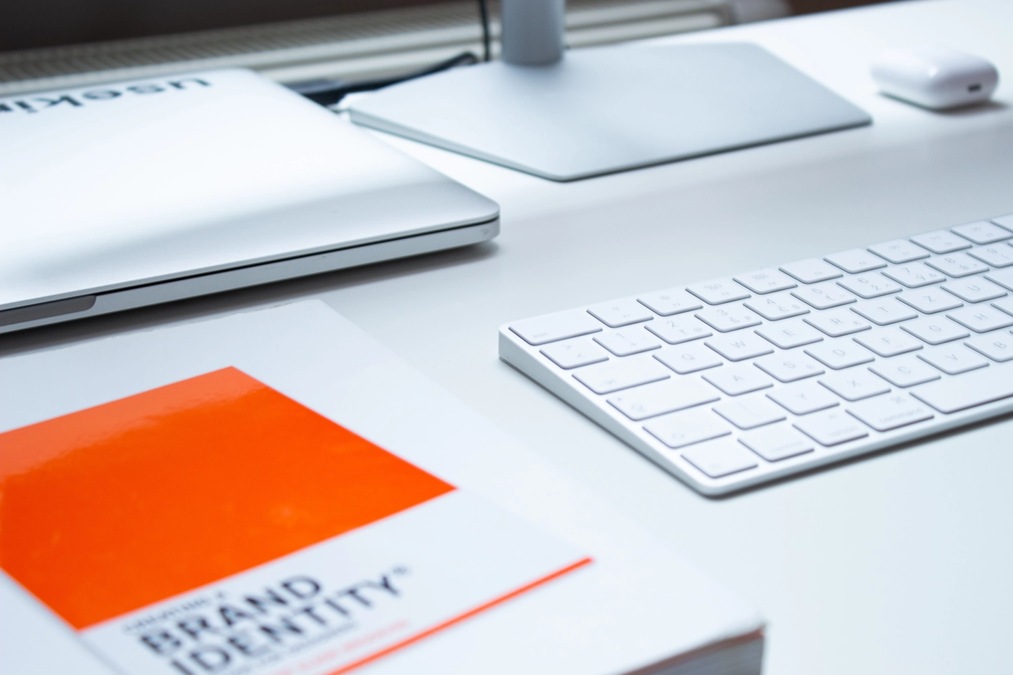Modernes Arbeitsplatz-Setup einer Mediengestalter:in mit einem Buch über Markenidentität auf dem Tisch neben einer weißen Tastatur und einem Laptop.